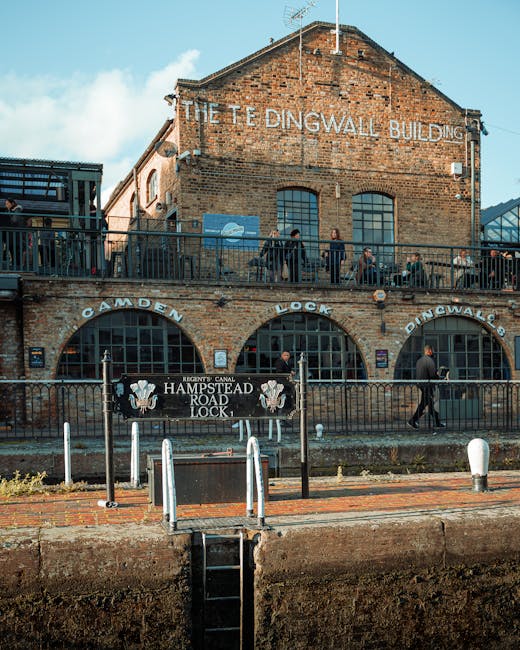 A photograph of the exterior of The Teddington Lock building, a brick structure with large arched windows on the ground floor and smaller rectangular windows above. The building has a sign indicating 'The Teddington Lock Building' at the top, with the words faded yet still visible. In front, there is a metal railing with signs labeled 'Camden Lock' and 'Teddington Lock,' along with a black banner reading 'Hampstead Road Lock,' featuring an illustrated emblem. Several people are walking on the sidewalk and along the upper balcony, some carrying bags or backpacks. The scene is captured during daytime with bright natural lighting, showing a lively area near the canal or river, with a pathway and bollards along the waterfront in the foreground. The image appears to be part of content related to house removals or relocation logistics, as it displays the setting associated with moving services.
