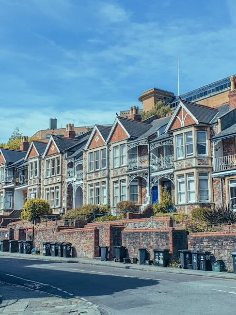An exterior view of a row of Victorian-style terraced houses in West Hampstead, featuring decorative ironwork balconies, bay windows, and brick facades. The houses are situated on a slightly elevated street, with a brick retaining wall separating them from the pavement. The street below has multiple black waste bins lined up along the curb. The sky above is clear with a few scattered clouds, indicating daytime. The image captures a residential area suitable for home relocation and moving services, with no visible moving activity or equipment present. An understanding of such environments is important for furniture transport and packing and moving processes, as provided by Removal Van West Hampstead.
