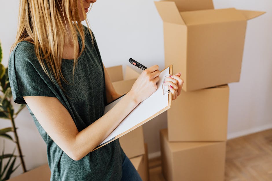 A woman with long blonde hair wearing a dark grey t-shirt is standing indoors, writing on a small white notepad with a black marker. In front of her, there are three stacked cardboard moving boxes, one of which is open with flaps raised, showing some empty interior space. The environment appears to be a home setting with a white wall and a potted plant visible in the background. The floor is made of wooden planks. The woman is engaged in a packing or planning task as part of a home relocation process, with the boxes likely prepared for moving day. The scene is well-lit with natural light, reflecting a typical preparatory phase of packing and furniture transport associated with house removals handled by companies like Removal Van West Hampstead, focusing on the essentials of packing lists and organising possessions for efficient loading and transport.