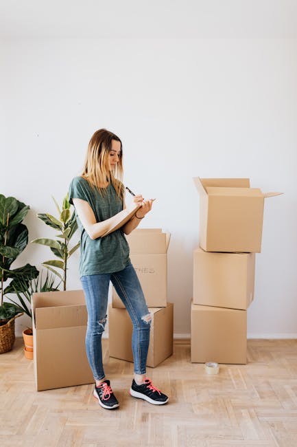 A young woman with shoulder-length blonde hair, dressed in a green t-shirt and blue ripped jeans, stands in a well-lit room with a white wall and wooden floor. She is holding a clipboard and pen, appearing to be inspecting or taking notes. Behind her are several cardboard moving boxes, some stacked and others open, with packing materials such as plastic wrap and paper inside. A large potted green plant with broad leaves is positioned to her left, adding a touch of greenery to the scene. The boxes are arranged near a doorway or an open space within the home, indicating an ongoing packing or home relocation process. The setting reflects a house removal preparatory stage, involving furniture transport, packing, and organizing belongings before a move, as handled by Removal Van West Hampstead.