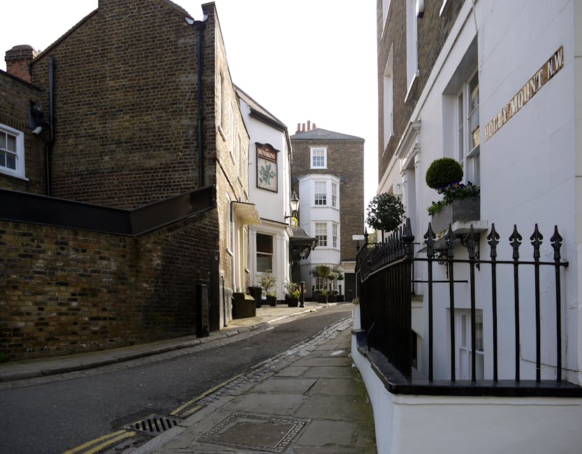 A three-storey red brick building on a narrow street with limited parking, featuring a ground-floor shop with green and white signage and large front windows. The upper floors have multiple white-fram
