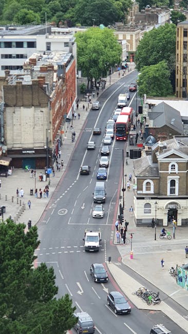 An aerial view of a busy urban street in West Hampstead NW6, showing multiple vehicles including cars, vans, and buses traveling along a winding road. On either side of the street, there are buildings