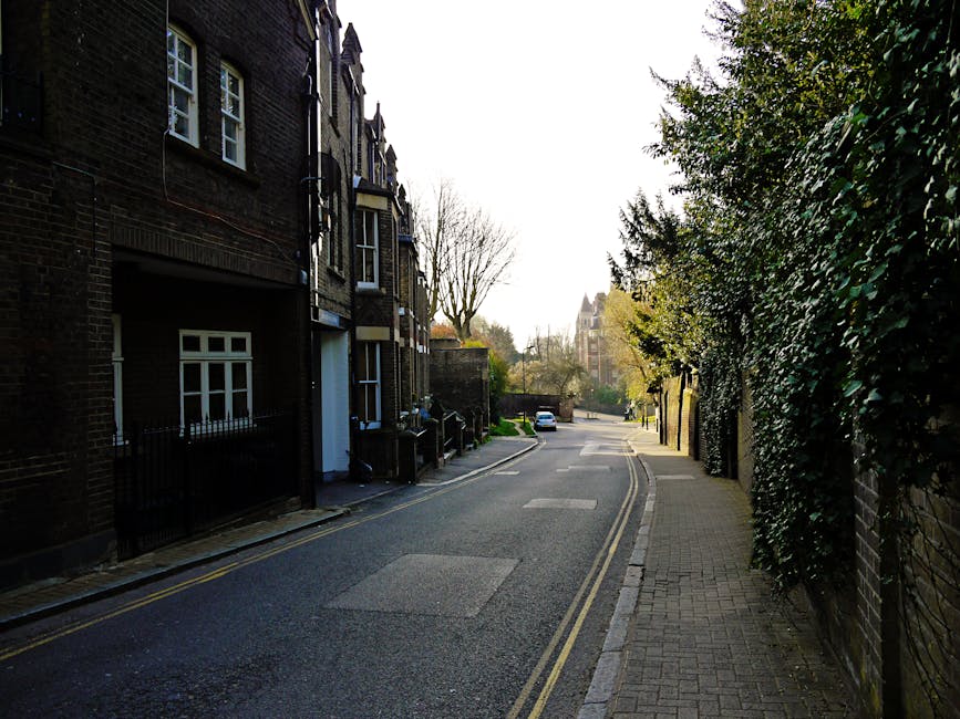 A quiet residential street in West Hampstead during daylight, featuring a narrow road with a double yellow line along the curbside. On the left, there are brick residential buildings with varying wind