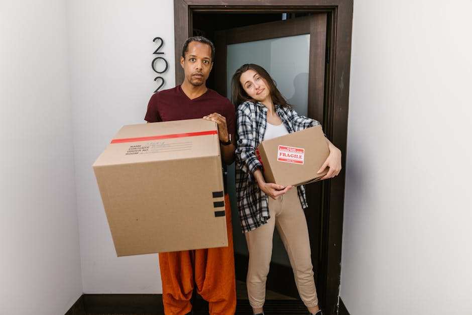 A man and a woman standing in a doorway inside a property, both holding cardboard boxes used for home relocation, with the man holding a larger box and the woman holding a smaller one labeled 'Fragile.' The man is dressed in a maroon t-shirt and orange trousers, while the woman wears a plaid shirt over a white top and beige trousers. Inside the doorway, visible behind them, are a dark wooden doorframe and a light-colored wall with the number '22' mounted vertically near the door frame. The scene captures the loading process of furniture transport and packing materials, with cardboard boxes ready for loading onto a removal van by Removal Van West Hampstead, as part of a professional house removals or moving service, illustrating the packing and moving stage of a home relocation in West Hampstead. Lighting is natural and evenly distributed, emphasizing the boxes and the individuals involved in the moving logistics.