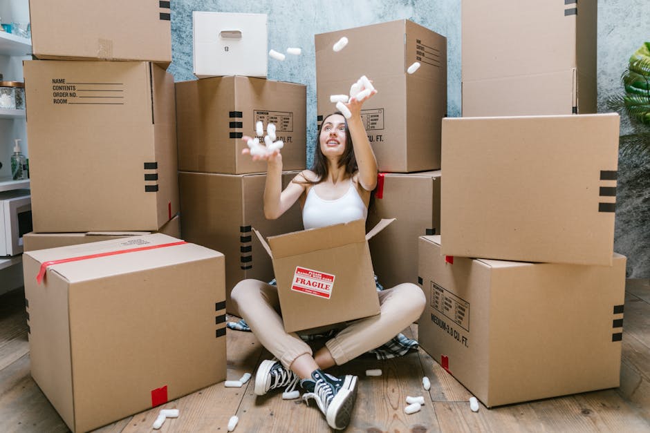 A young woman sits cross-legged on a wooden floor inside a residential room, surrounded by stacked cardboard boxes used for home relocation, some sealed with red tape and others open. She is smiling while tossing packing foam peanuts into the air, with additional foam peanuts spilling from an open box in her lap. The boxes vary in size, with labels indicating contents and fragile warnings visible on some. The background shows light blue textured walls and a small potted plant to the right, with part of a white kitchen countertop and cupboard visible in the left corner. The scene depicts the packing and moving process, highlighting the careful handling of household items, with no furniture or appliances in view. This image, associated with Removal Van West Hampstead, illustrates the preparation phase of furniture transport during a house move, emphasizing organized packing and logistical planning.