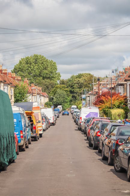 A quiet residential street in West Hampstead NW6 with rows of parked cars on both sides, including hatchbacks, saloons, and vans. The street is paved with asphalt, and there are several houses with brick and stucco facades, some featuring small front gardens with bushes and trees. Overhead, a network of electrical wires stretches across the sky, which is partly cloudy with patches of blue. In the background, larger green trees rise above the rooftops, indicating a green, suburban environment. This scene may depict a typical day before a house removal or furniture transport, with the possibility of a Removal Van West Hampstead team preparing to load items into a van parked nearby, utilizing trolleys, straps, or blankets for safe moving and packing during a home relocation process.