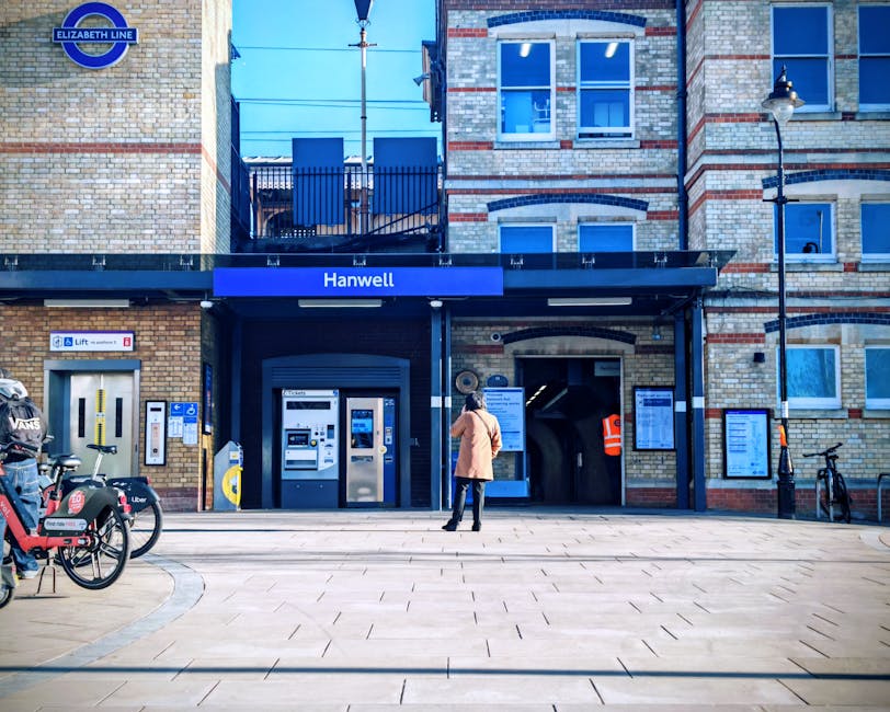 A woman in a beige coat and black trousers is standing outside Hanwell station entrance, which features a blue sign and a modern brick facade with white window frames. She appears to be reading an information board or timetable mounted on the wall beside the station entrance. In front of her, on the left side of the image, there are bicycles parked, branded with Uber and VANS logos, secured with bike locks. To the right of the station entrance, there is a small bicycle rack and a black trash bin. The station's entrance includes ticket machines and a lift for accessible access, with a pathway paved with light-colored tiles leading up to the station doors. A pedestrian stands on the pavement near the entrance, dressed for cold weather. The environment is well-lit with natural daylight, and a street lamp is mounted on the building's exterior wall. This scene exemplifies the kind of urban setting where Removal Van West Hampstead might facilitate home relocation or furniture transport services, especially in areas with accessible public transportation like Hanwell station.