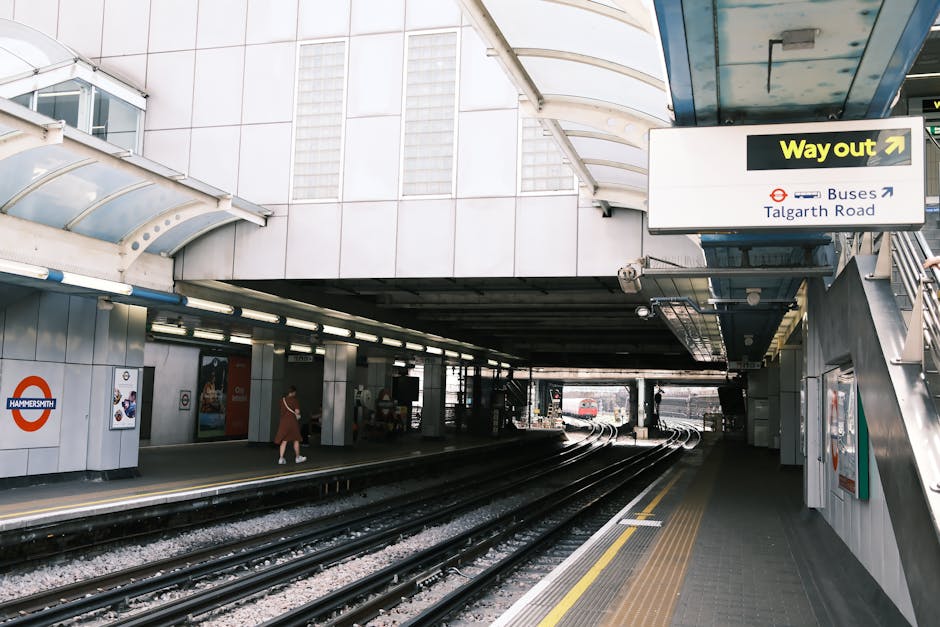 A view of West Hampstead station entrance and approach, showing train tracks running underneath a canopy with an overhang supported by metal beams. To the left, there are white tiled walls with station signage, including a roundel sign for Hammersmith, and a woman in a beige coat walking along the platform. Overhead, a large black and yellow sign indicates the way to the bus station and Talgarth Road, with an arrow pointing right. On the stairway leading to the station exit, an escalator or staircase is partially visible, and the platform area appears prepared for passenger movement and home relocation tasks. The environment is well-lit, with natural light filtering through the canopy and ambient lighting illuminating the area, making it suitable for loading moving boxes, furniture, or appliances during a furniture transport or home removal process by Removal Van West Hampstead.