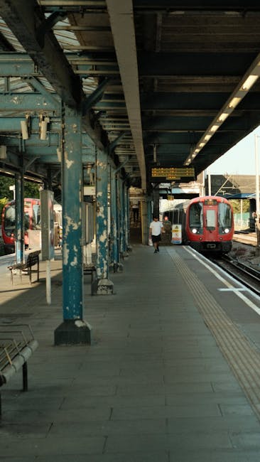 A woman in a beige coat and black trousers is standing outside Hanwell station entrance, which features a blue sign and a modern brick facade with white window frames. She appears to be reading an information board or timetable mounted on the wall beside the station entrance. In front of her, on the left side of the image, there are bicycles parked, branded with Uber and VANS logos, secured with bike locks. To the right of the station entrance, there is a small bicycle rack and a black trash bin. The station's entrance includes ticket machines and a lift for accessible access, with a pathway paved with light-colored tiles leading up to the station doors. A pedestrian stands on the pavement near the entrance, dressed for cold weather. The environment is well-lit with natural daylight, and a street lamp is mounted on the building's exterior wall. This scene exemplifies the kind of urban setting where Removal Van West Hampstead might facilitate home relocation or furniture transport services, especially in areas with accessible public transportation like Hanwell station.